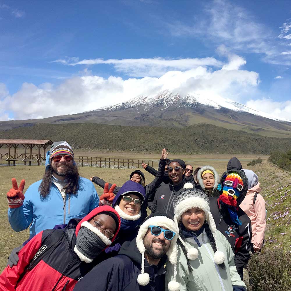 a group of warmly dressed students smiling and waving as a volcano billows smoke into the blue sky
